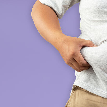 Bildausschnitt einer Person in T-Shirt und Hose vor hell-violettem Hintergrund, die sich mit einer Hand in den Bauch kneift.