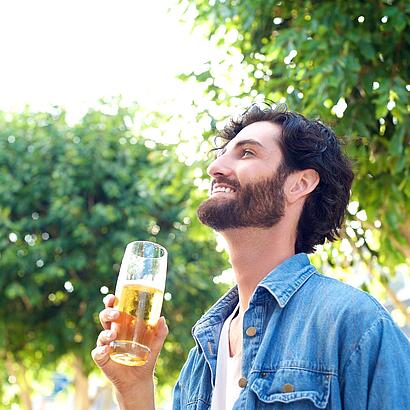Ein Mann trinkt ein Glas Bier und schaut fröhlich nach oben. Im Hintergrund sind grüne Bäume und ein sonniger Himmel.