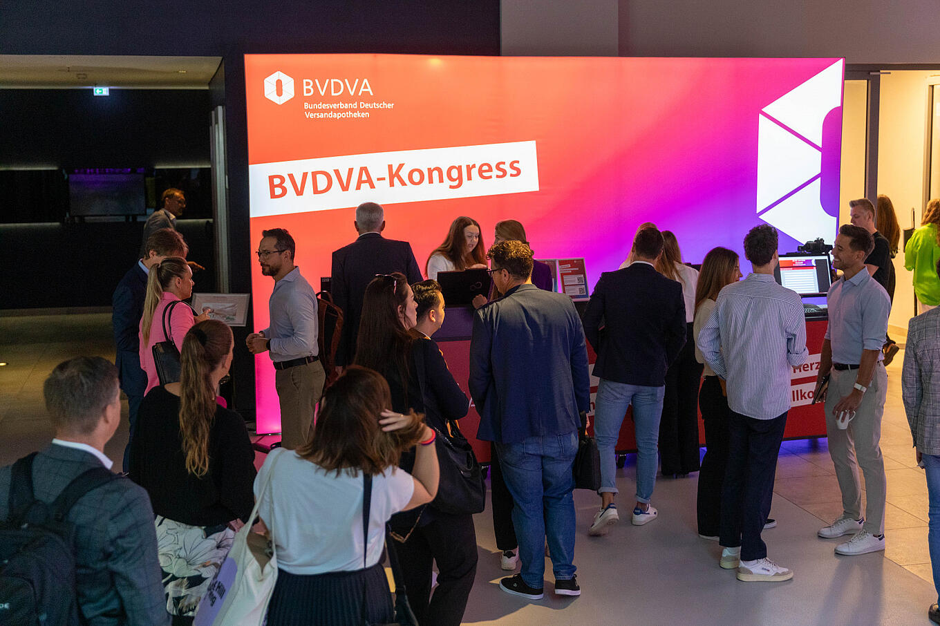 Vor einem Wanddisplay mit der Aufschrift BVDVA-Kongress stehen Menschen an einem Tresen mit Bildschirmen.