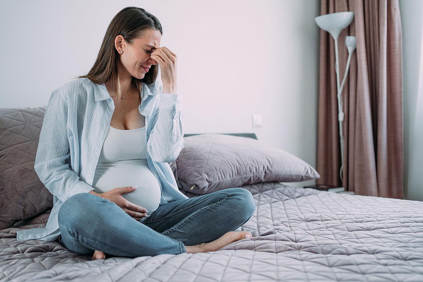 Schwangere Frau mit Kopfschmerzen auf dem Bett sitzend