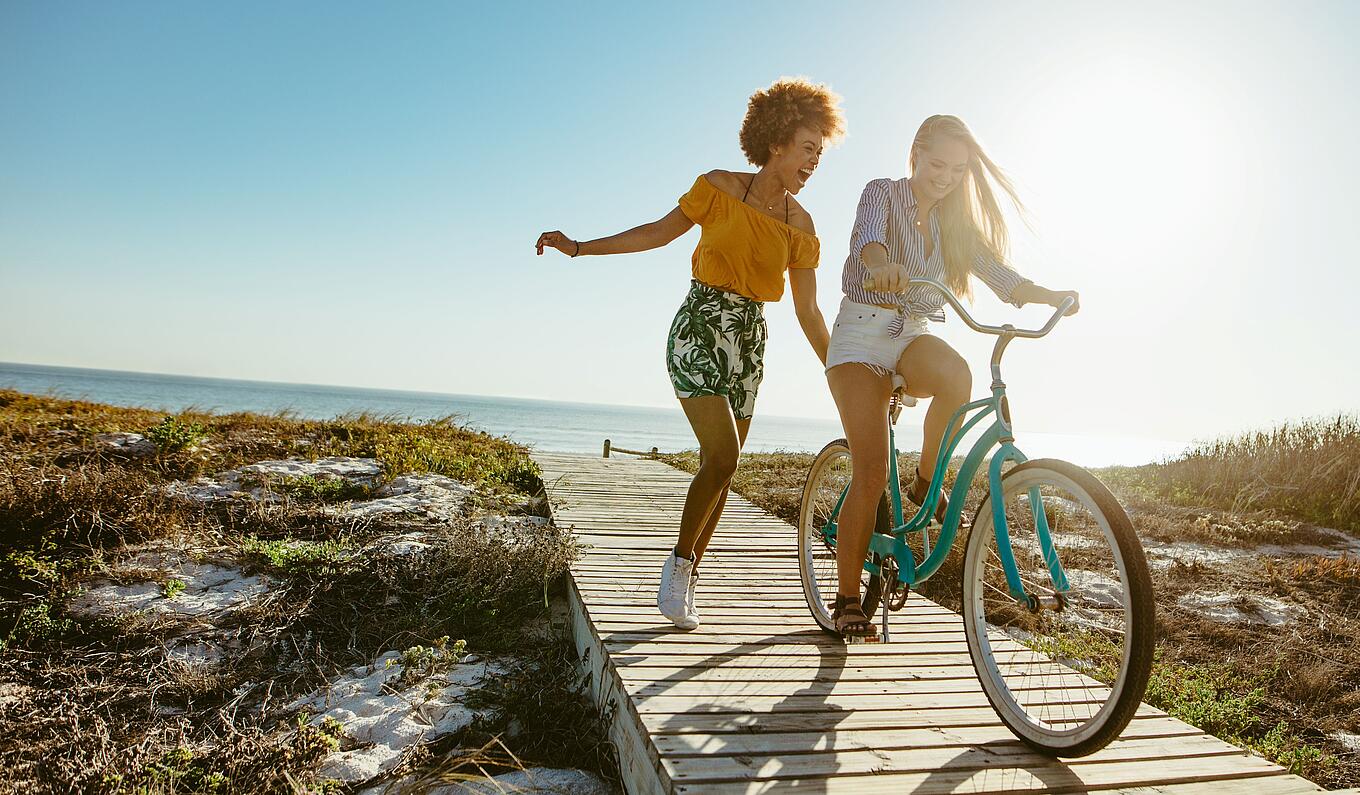 Sommerbild mit einer Frau auf dem Fahrrad, die andere Frau läuft unbeschwert nebenher auf einem Steg