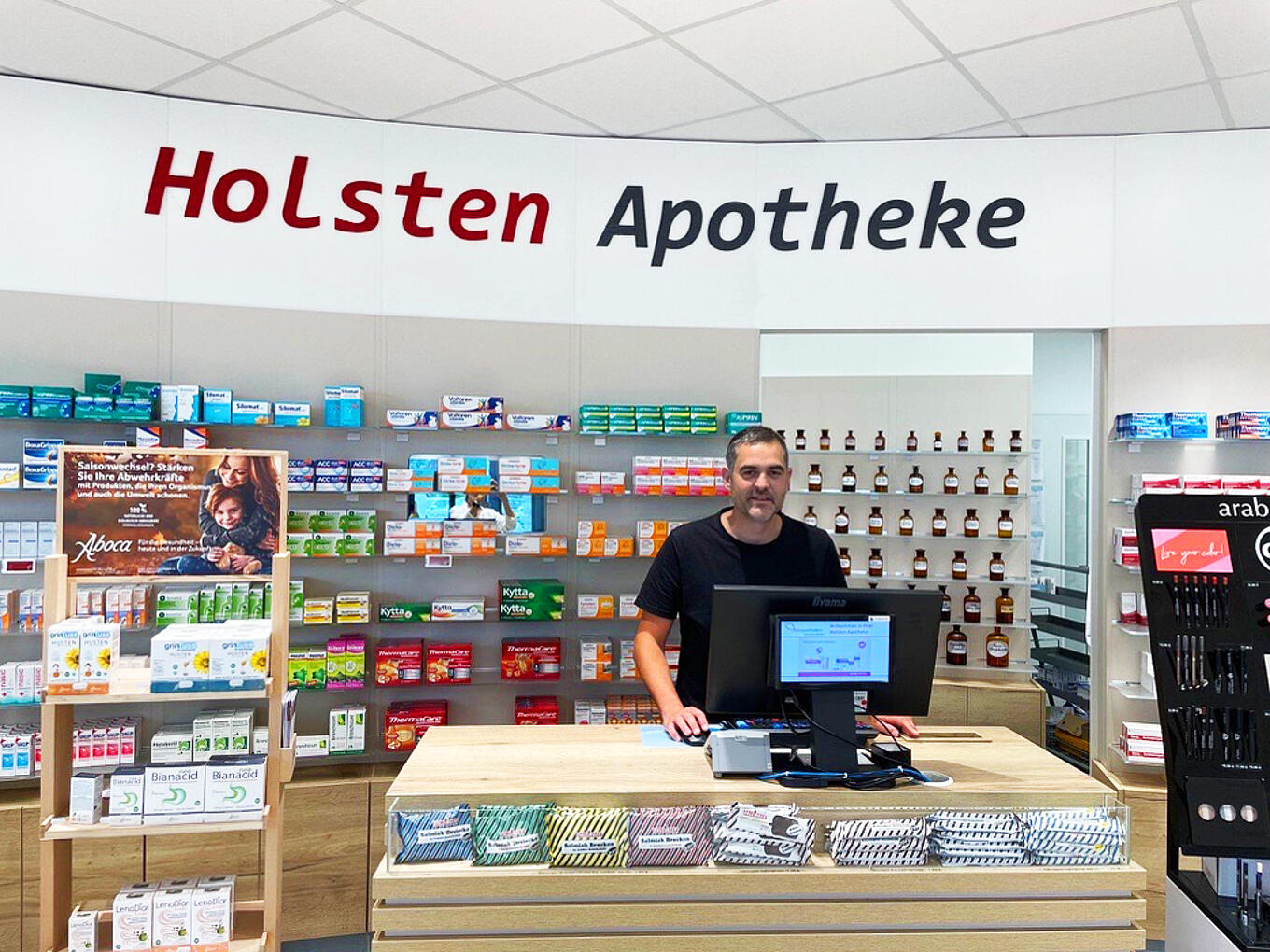 Ein Mann steht hinter dem Tresen in einer Apotheke. Über ihm befindet sich die Aufschrift Holsten Apotheke.