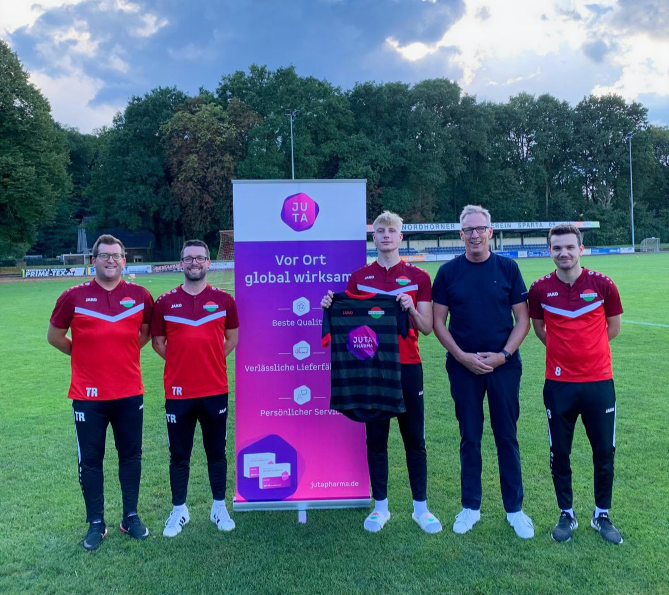 Bild von Fußballspielern und Vertriebsleiter mit einem Tricott und dem Juta Logo sowie einem Banneraufsteller von Juta Pharma.