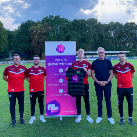 Bild von Fußballspielern und Vertriebsleiter mit einem Tricott und dem Juta Logo sowie einem Banneraufsteller von Juta Pharma.